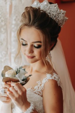 Güzel elbiseli gelin, elinde damadın çiçeğiyle pencerenin yanında duruyor. Düğün için. Düğün buketi Gelinin Sabahı. Mutlu bir gelin damadı bekliyor..