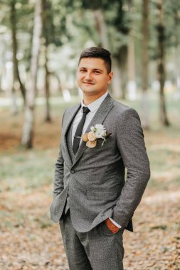 Süslü bir damat ormanda yürürken ceketini uzun ağaçların arasına ilikler. Modaya uygun bir damat, yetişkin bir adam.