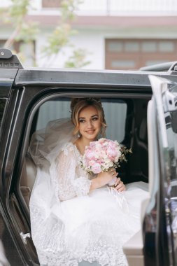 Sarışın bir gelin düğün gününde siyah bir arabada bir buketle oturur. Gelinin portresi. Güzel makyaj ve saç. Muhteşem beyaz dantel elbise