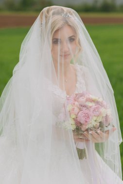 Gelinin portresi. Gelin, yeşil bir tarlada duvak ile kaplı beyaz bir elbise içinde bir buketle duruyor. Harika bir elbise. Özel bir kadın. Düğün fotoğrafı.