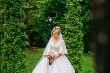 Gelinin geniş portresi. Gelin, yemyeşil ağaçların arka planında beyaz bir elbise ve bir buket ile duruyor. Harika bir elbise. Özel bir kadın. Düğün fotoğrafı.