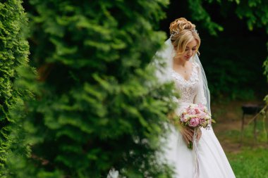 Gelinin geniş portresi. Gelin, yemyeşil ağaçların arka planında beyaz bir elbise ve bir buket ile duruyor. Harika bir elbise. Özel bir kadın. Düğün fotoğrafı.