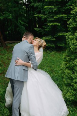 Gelin ve damadın portresi yeşil ağaçların arkasında duruyor, damat gelinin alyansını takıyor. Şık damat. Moda ve tarz.