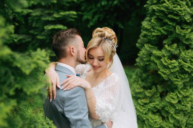 Gelin ve damadın portresi yeşil ağaçların arka planında duruyor, kucaklıyor. Gelin içtenlikle gülümsüyor. Şık damat. Moda ve tarz. Güzel gelin.