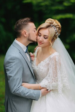 Gelin ve damadın portresi yeşil ağaçların arkasında duruyor, damat gelinin alyansını takıyor. Şık damat. Moda ve tarz.