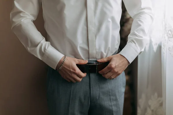 Details. A man in a white shirt and gray pants adjusts his stylish belt. Masculine style. Fashion. Business