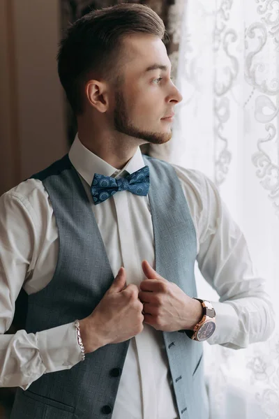A man in a white shirt and a gray vest poses by the window and looks to the side, adjusts his vest. Front view. A stylish watch. Men\'s style. Fashion. Business