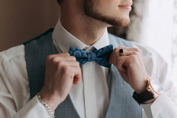 白いシャツにグレーのベストを着た男が窓からポーズをとって横を見ると 青い弓のネクタイが調整されています 正面図 スタイリッシュな時計です 男性のスタイル ファッションだ — ストック写真