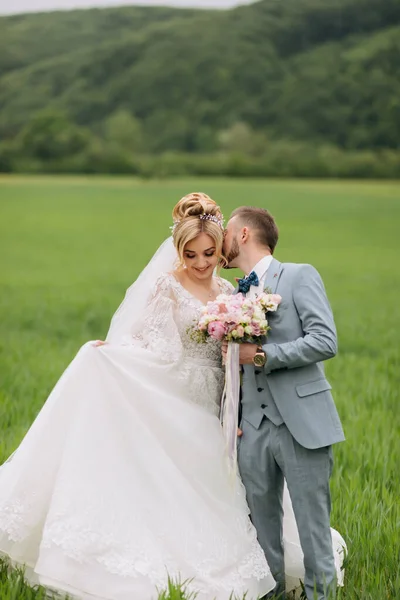新婚夫婦は緑の畑に立ってキス 花嫁は花束を持っている 自然の中で結婚式 花嫁と新郎の肖像画 スタイリッシュな新郎 — ストック写真
