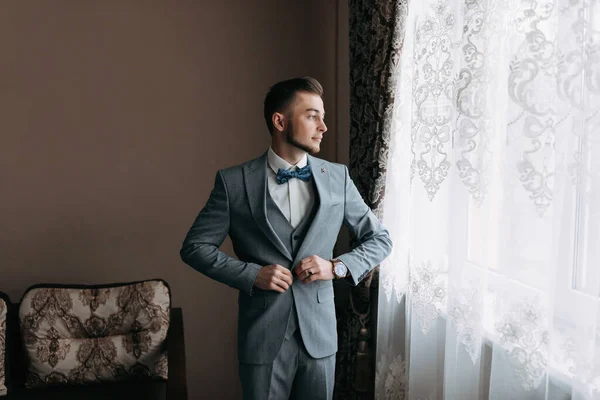 一个穿着白衬衫和灰色西服的男人站在窗前 把目光移开 调整他的外套 前面的景色一个时髦的手表 男人的风格 — 图库照片