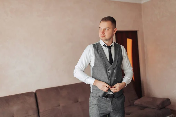 一个穿着白衬衫和灰色背心的男人站在窗边看镜头 调整背心 前面的景色一个时髦的手表 男人的风格 — 图库照片