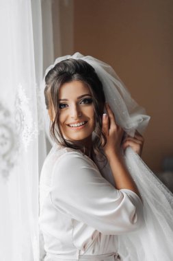 Gelinin portresi. Sabahlık giymiş esmer bir gelin, poz veriyor, şehvetli beyaz duvağını tutuyor ve kamera lensine bakıyor. Harika bir makyaj ve saç. Düğün fotoğrafı. Güzel gelin.
