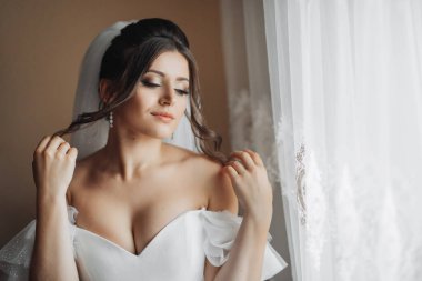 Beyaz elbiseli esmer bir gelin muhteşem buklelerini tutar ve aşağı bakar. Omuzlarını aç. Güzel eller. Uzun peçe. Gelinin sabahı.