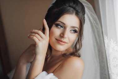 Beyaz elbiseli esmer bir gelin muhteşem buklelerini tutar ve aşağı bakar. Omuzlarını aç. Güzel eller. Uzun peçe. Gelinin sabahı.