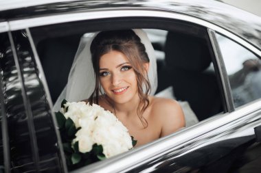 Esmer bir gelin, tüm samimiyetiyle gülümseyerek düğün gününde siyah bir arabada bir buket beyaz gülle oturur. Gelinin portresi. Güzel bukleler. Güzel makyaj ve saç. Lüks beyaz bir elbise.