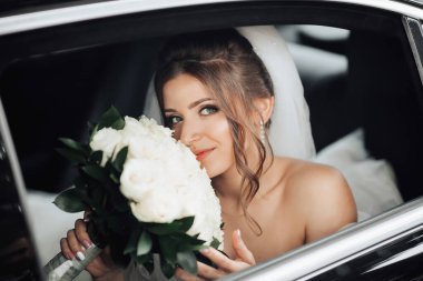 Esmer bir gelin, tüm samimiyetiyle gülümseyerek düğün gününde siyah bir arabada bir buket beyaz gülle oturur. Gelinin portresi. Güzel bukleler. Güzel makyaj ve saç. Lüks beyaz bir elbise.