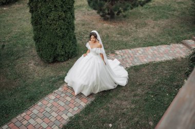 Düğün fotoğrafı. Şehvetli beyaz bir elbise ve uzun bir peçe ile gelin taş bir patika boyunca bahçede yürür. Yukarıdan bak. Gelinin portresi. Güzel bukleler. Güzel makyaj ve saç.
