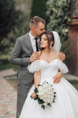 Gelin ve damadın doğadaki portresi. Damat geline arkadan sarılır ve onu öper, beyaz elbiseli gelin bir buket beyaz gül tutar ve yüzünü çevirir. Güzel makyaj ve saç.