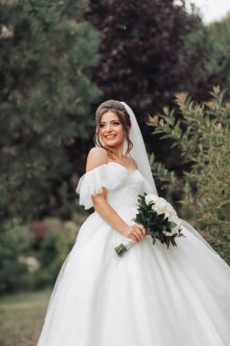 Düğün fotoğrafı. Şehvetli beyaz bir elbise ve uzun bir duvak takmış, gülümseyen, elinde bir buket beyaz gülle elbisesini tutan gelin. Gelinin portresi. Güzel makyaj ve saç.