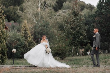 Gelin bir buket gül tutuyor, damadın önünde yürüyor ve ona omuzlarının arkasından bakıyor. Geniş açılı fotoğraf. Portre. Doğada düğün. Güzel saç ve makyaj. Şık damat