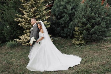 Gelin ve damadın doğadaki portresi. Gelin ve damat, kozalaklı ağaçların arka planında ayakta, kucaklaşıyorlar. Bir buket güllü, uzun elbiseli gelin. Şık damat.