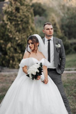 Gelin ve damadın doğadaki portresi. Damat gelinin arkasında duruyor, kozalaklı ağaçların arkasında poz veriyor. Bir buket güllü, uzun elbiseli gelin. Şık damat.