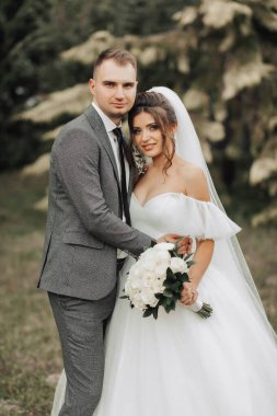 Gelin ve damadın doğadaki portresi. Kesilmiş fotoğraf. Gelin ve damat, kozalaklı ağaçların arkasına sarılıp gülümsüyor. Bir buket gül taşıyan uzun elbiseli gelin..