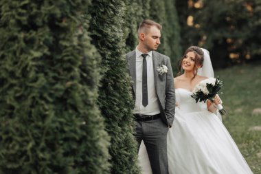 Gelin ve damadın doğadaki portresi. Gri takım elbiseli damat ön planda, gelin arkasında yürüyor, elinde bir buket buketle, beyaz, şehvetli bir elbise içinde gülümsüyor. Şık damat