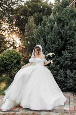 Doğadaki gelinin portresi. Şehvetli bir elbise ve duvak içinde esmer bir gelin, ayakta, bir buketle ormanın arka planına ve ahşap bir kulübeye karşı poz veriyor. Güneşin güzel bir ışığı.