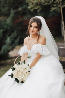 Doğadaki gelinin portresi. Beyaz uzun elbiseli esmer gelin, elinde bir buket beyaz gül, bir bankta oturuyor, poz veriyor, aşağı bakıyor, ağaçların arkasına bakıyor. Kıvırcık saçlı