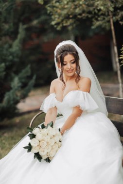 Doğadaki gelinin portresi. Beyaz uzun elbiseli esmer gelin, elinde bir buket beyaz gül, bir bankta oturuyor, poz veriyor, aşağı bakıyor, ağaçların arkasına bakıyor. Kıvırcık saçlı