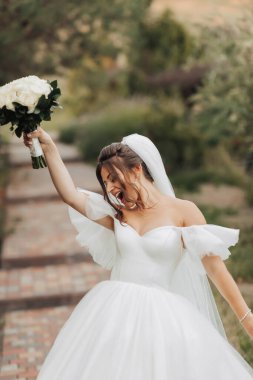 Doğadaki gelinin portresi. Beyaz uzun elbiseli esmer bir gelin, elinde bir buket beyaz gül, poz veriyor, objektife bakıyor, içtenlikle gülümsüyor. Ağacın arka planında. Kıvırcık saçlı.