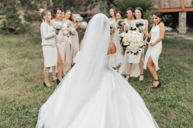 Doğada düğün fotoğrafı. Uzun beyaz elbiseli esmer bir gelin çıplak elbiseli arkadaşlarının önünde duruyor. Gülümsüyorlar ve elinde Gypila buketleri var. Genç kadınlar. duygular