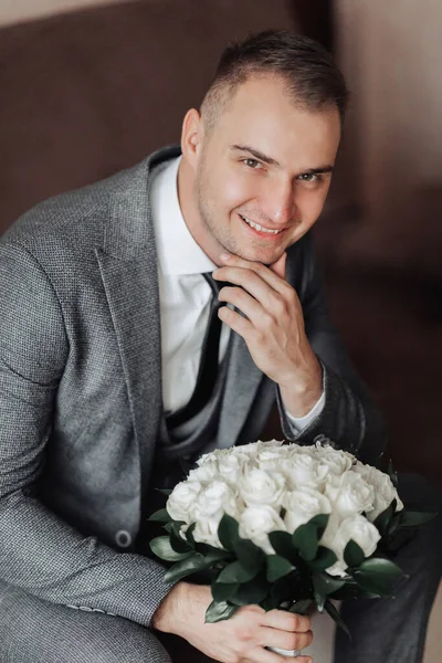 肖像画 白いシャツ 黒いネクタイ 灰色のスーツの男がソファに座って 白いバラの花束を持っています スタイリッシュな時計です 男性のスタイル ファッションだ — ストック写真