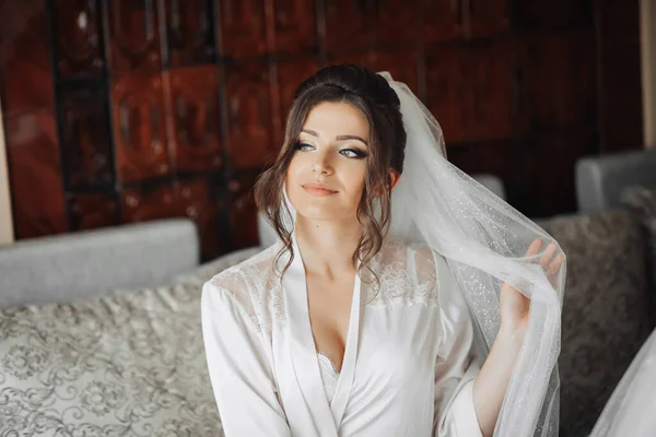 新娘的画像 一位黑发新娘穿着睡衣坐在灰色的沙发上 摆出一副长长的白色面纱 漂亮的妆容和头发婚纱照漂亮的新娘 — 图库照片
