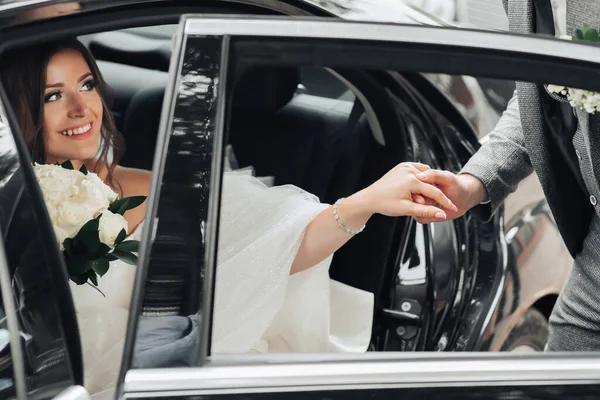 婚礼当天 一位黑发新娘正笑着坐在一辆黑色的车上 与新郎握手 看着新郎 新娘的画像 漂亮的妆容和头发一件豪华的白色连衣裙 — 图库照片