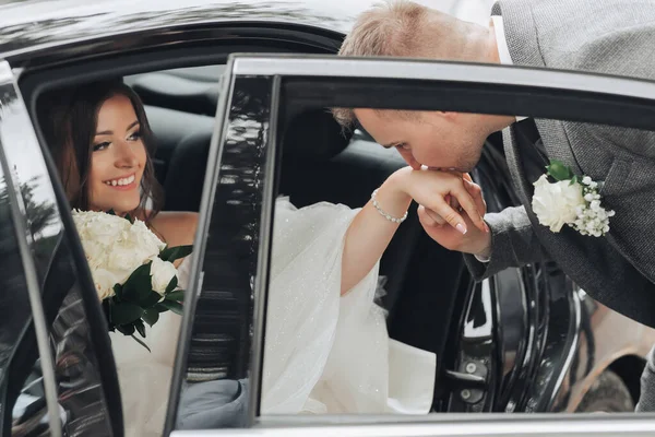 婚纱照新娘笑着坐在一辆黑色的车上 新郎亲吻新娘的手 从车窗往外看 漂亮的妆容和头发一件豪华的白色连衣裙 — 图库照片