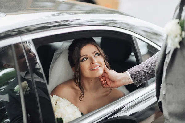 結婚式の写真 花嫁は笑顔で黒い車の中に座って 新郎は優しく花嫁のあごを保持します 車の窓からの眺め 美しいメイクや髪 豪華な白いドレス — ストック写真