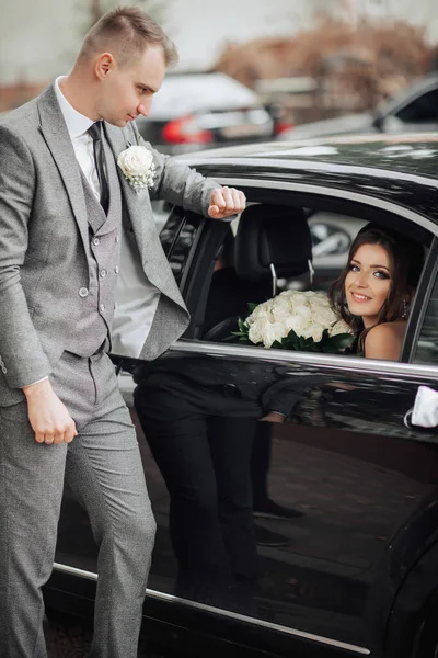 婚纱照新娘拿着一束玫瑰坐在一辆黑色的车上笑着 新郎靠在车上看着新娘 漂亮的妆容和头发一个时髦的男人 — 图库照片
