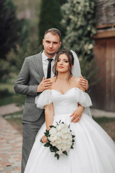 Portrait of the bride and groom in nature. The groom hugs the bride from behind, the bride in a white dress holds a bouquet of white roses and looks at the camera. Beautiful makeup and hair.