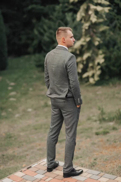灰色のスーツの男はポケットに手を入れて自然にポーズをとり 肩はカメラに向かった 後ろの景色 スタイリッシュな時計です 男性のスタイル ファッションだ — ストック写真
