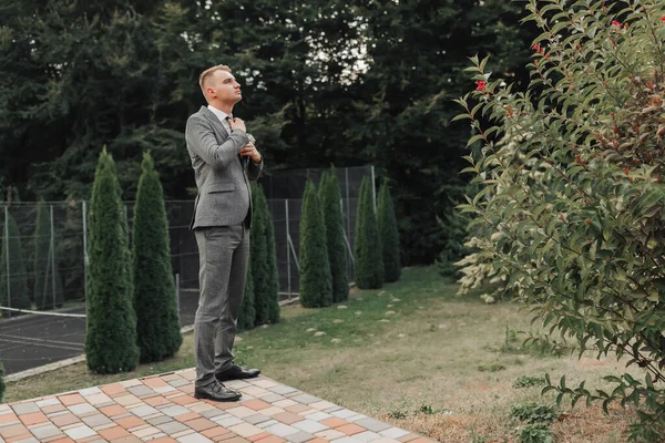 一个穿着灰色西装的男人在自然中摆姿势 调整领带 侧视图 一个时髦的手表 男人的风格 — 图库照片