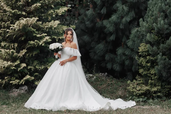 ボリュームのある白いドレスと長いベールの花嫁は 緑の背景に立って白いバラの花束を持ち 彼女のドレスを保持しています 花嫁の肖像画 美しいメイクと髪 — ストック写真