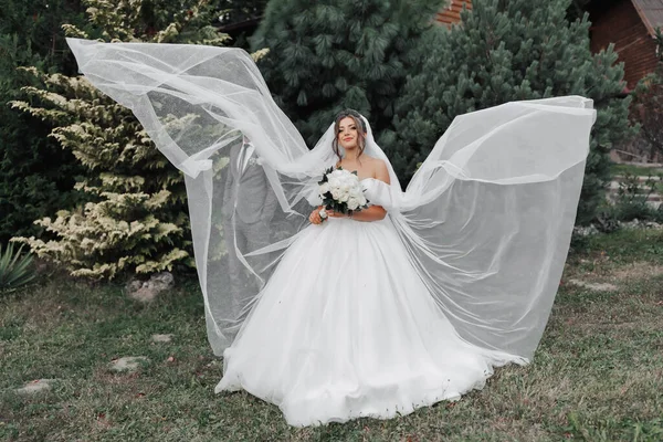 白いドレスとベールの中髪のブルネットの花嫁は 空気中で美しく流れる 彼女のスタイリッシュな新郎の前に立っています 花嫁と新郎の肖像画 自然界の結婚式 — ストック写真