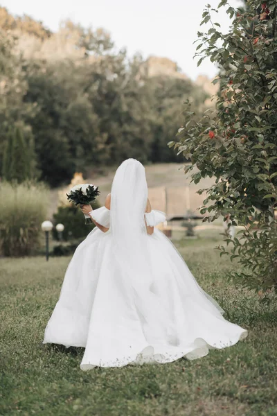 婚纱照新娘身穿厚重的白色连衣裙 头戴长长的面纱 走在石子路旁的花园里 手里拿着一束白玫瑰 背对着摄像机 — 图库照片