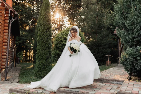 花嫁の肖像画です ボリュームのあるドレスとベールの中にあるブルネットの花嫁は 森と木製の小屋の背景に花束でポーズをとって立っています 美しい太陽の光 — ストック写真