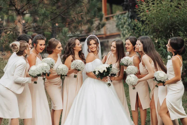 自然の中で結婚式の写真 白い長いドレスを着たブルネットの花嫁とヌードドレスの彼女の友人は 木の背景に立って 笑みを浮かべて ジプシーの花束を持っています 若い女性 — ストック写真