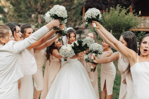 stock image Wedding photo in nature. A brunette bride in a white long dress and her friends in nude dresses are standing against the background of trees, smiling, holding up their gypsophila bouquets. Young women