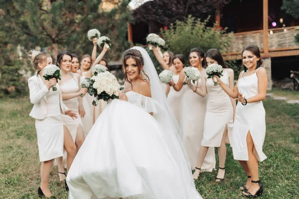 自然の中で結婚式の写真 長い白いドレスを着たブルネットの花嫁は 彼らのジプシーの花束を持ち上げ 笑顔で立っているヌードドレスの彼女の友人の前に立っています 若い女性だ — ストック写真