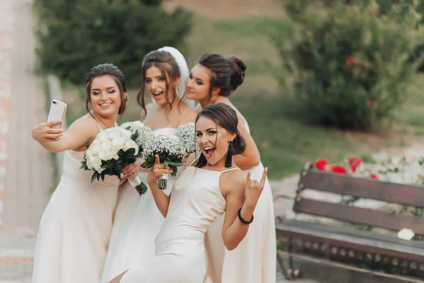 自然の中で結婚式の写真 長い白いドレスを着たブルネットの花嫁とヌードドレスの彼女の友人は笑顔で 自分の携帯電話で自撮りをして ジプシーの花束を持っています 若い女性だ — ストック写真